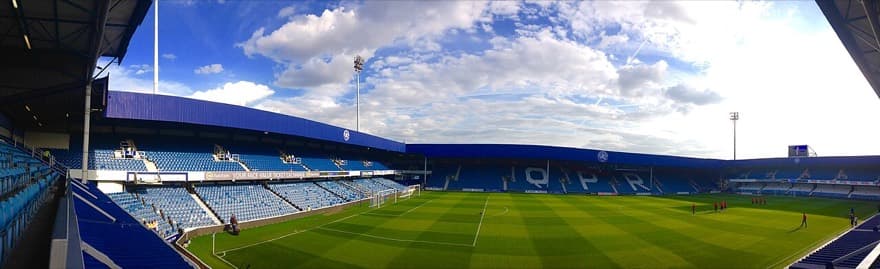 Loftus Road por dentro, com QPR escrito em branco nas cadeiras azuis da arquibancada oposta.